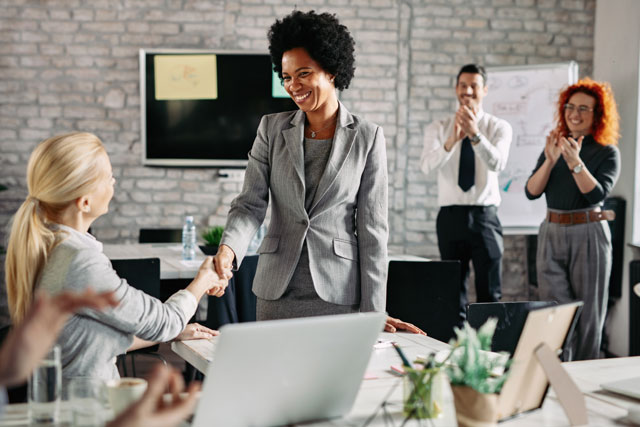 Happy businesswoman onboarding employee shaking hands with her colleague in the office while other co-workers are applauding them